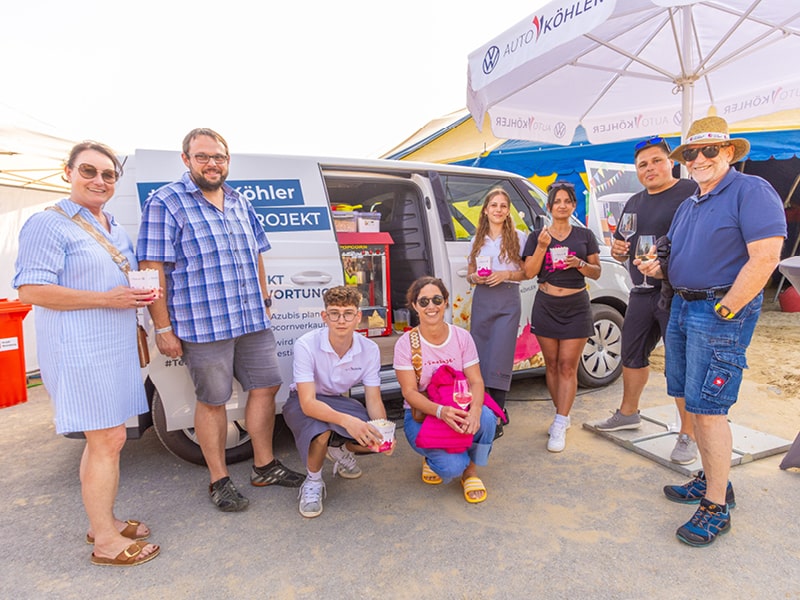 Zu sehen sind drei Personen des Caterer-Teams hinter einer aufgebauten Bar. Über ihnen prangt ein weißer Schriftzug auf blauem Hintergrund mit der Aufschrift: „Willkommen bei Volkswagen“