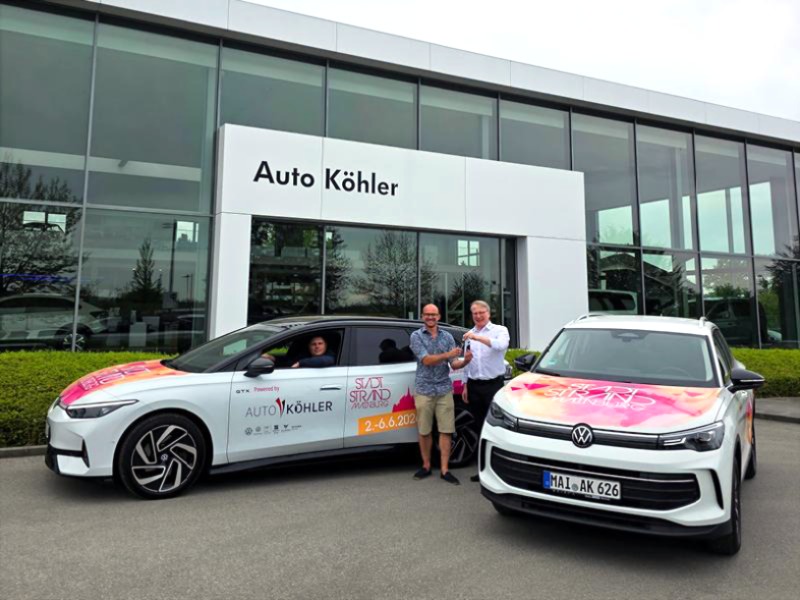 Zu sehen ist das Autohaus Auto Köhler, vor dem drei Männer stehen und symbolisch einen Autoschlüssel übergeben, während daneben zwei farbenfrohe beklebte VW-Fahrzeuge für das Event "Stadtstrand Wainburg 2024" werben.